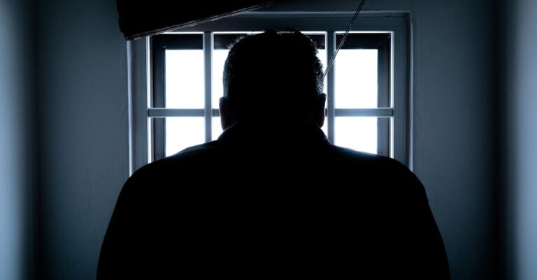 Dark silhouette of a man in a prison cell, backlit by window bars.